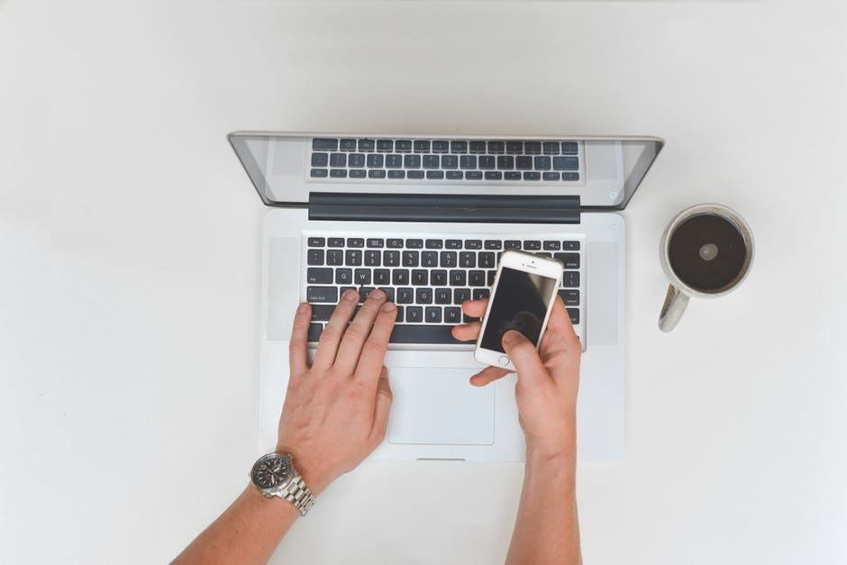 a person multitasks on a laptop while holding a smartphone in a modern workspace.