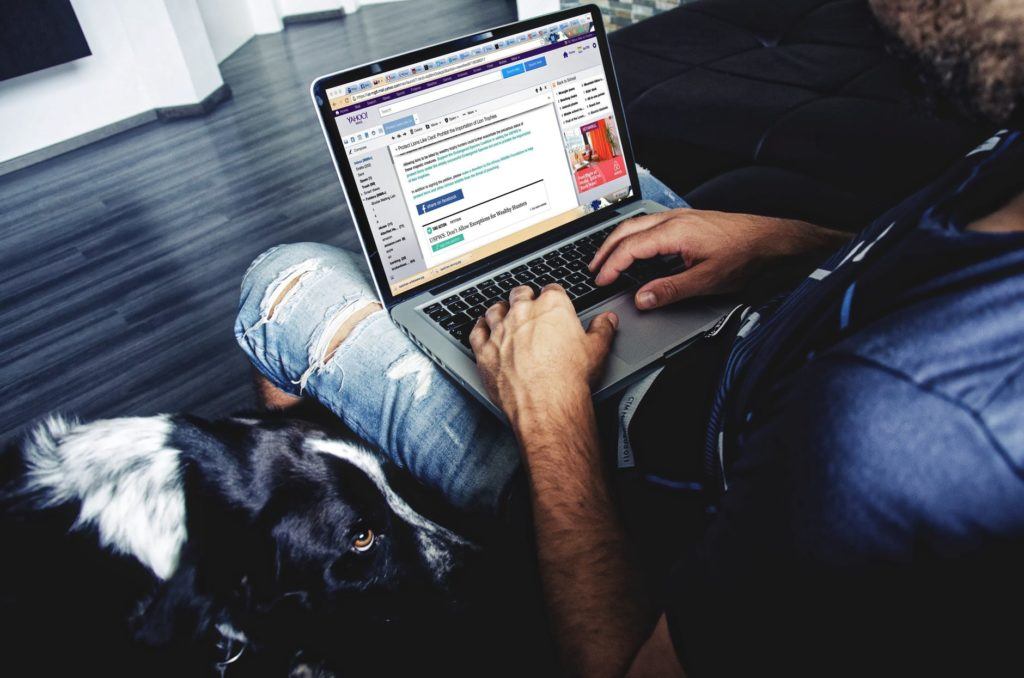 a man sitting on a couch using a laptop computer next to a dog