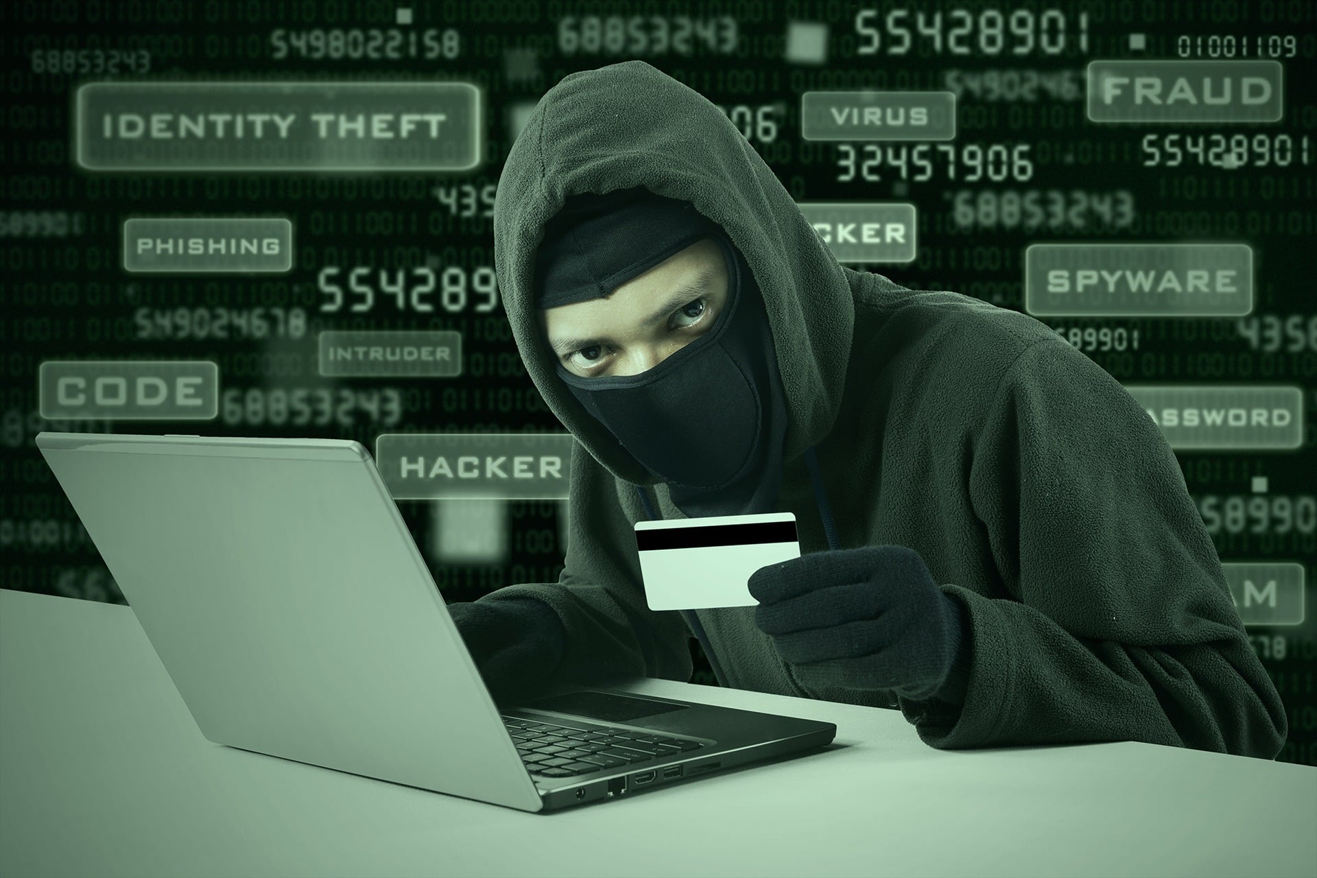 a man in a mask holding a credit card and looking at a laptop
